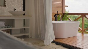 Un baño con un lavamanos blanco, una bañera grande y una terraza de madera con vistas a zonas verdes.