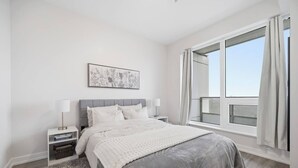 A modern bedroom with a grey bed, white bedding, a side table with a lamp, a framed picture on the wall, and a large window with curtains. - Contemporary Condo in DTK | Gym, Pool, & More (Kitchener)