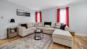 A living room with a sectional sofa, a small side table, a round coffee table, a rug, and a window with red curtains. - Spacious 3BR Upper Unit | Private Patio (Kitchener)