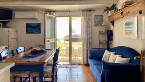 Une pièce comprenant une table à manger, des chaises, un canapé, un téléviseur et une porte donnant sur un espace extérieur agrémenté de plantes et offrant une vue.