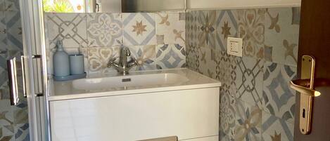 A bathroom with a white vanity, a mirror, patterned tiles, a towel, and a trash bin.