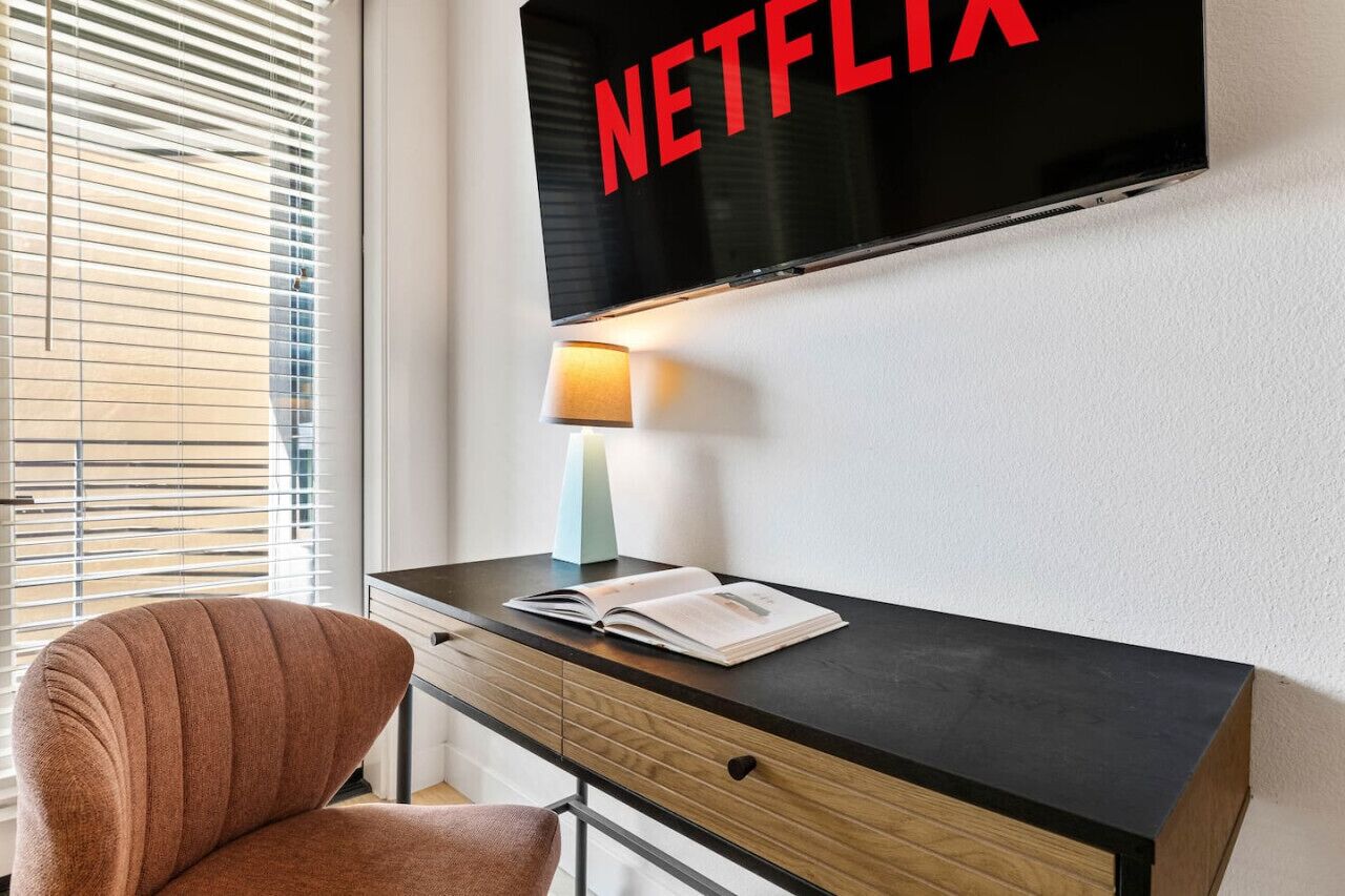 A modern room with a flat-screen TV mounted on the wall displaying 'NETFLIX,' a desk with a lamp, and a chair.