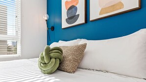 A bed with white bedding, a green decorative pillow, and two abstract paintings on a blue wall. - Bright & Modern Condo | 1Mi to South Coast Plaza (Santa Ana)