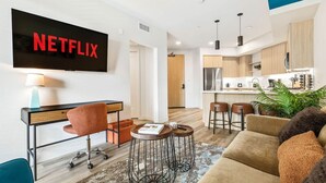 A modern living room with a TV, a desk, a sofa, and a kitchen in the background. - Bright & Modern Condo | 1Mi to South Coast Plaza (Santa Ana)