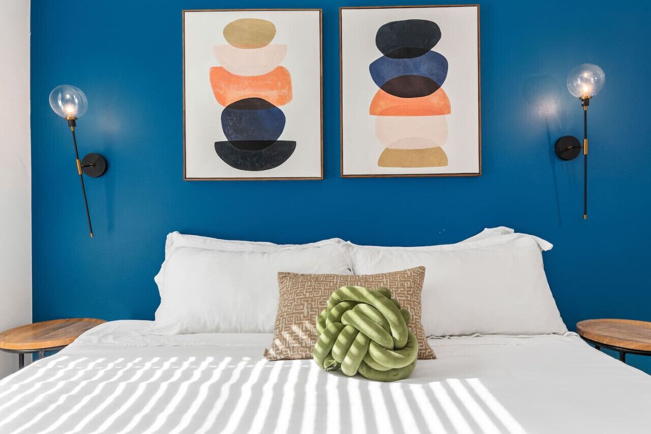 Una cama con ropa blanca, un almohadón verde y dos cuadros abstractos enmarcados sobre una pared azul.