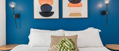 Una cama con ropa blanca, un almohadón verde y dos cuadros abstractos enmarcados sobre una pared azul.