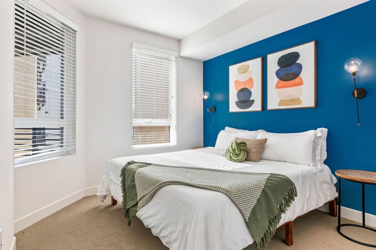 A bedroom with a bed, two windows with blinds, a blue accent wall, and two framed artworks.
