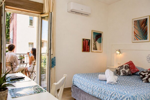 Una habitación con cama, una mesa, sillas, un balcón con vistas y dos cuadros en la pared.