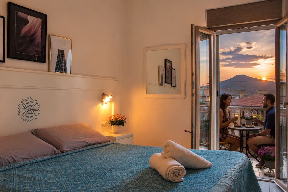 Une chambre à coucher avec un lit, une table de chevet, une lampe fixée au mur et une vue sur un balcon où deux personnes sont assises.