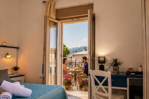 A room with a bed, a table with two people dining, a chair, a desk, and a view of the outside. - Inah Boutique Rooms (Dorgali)