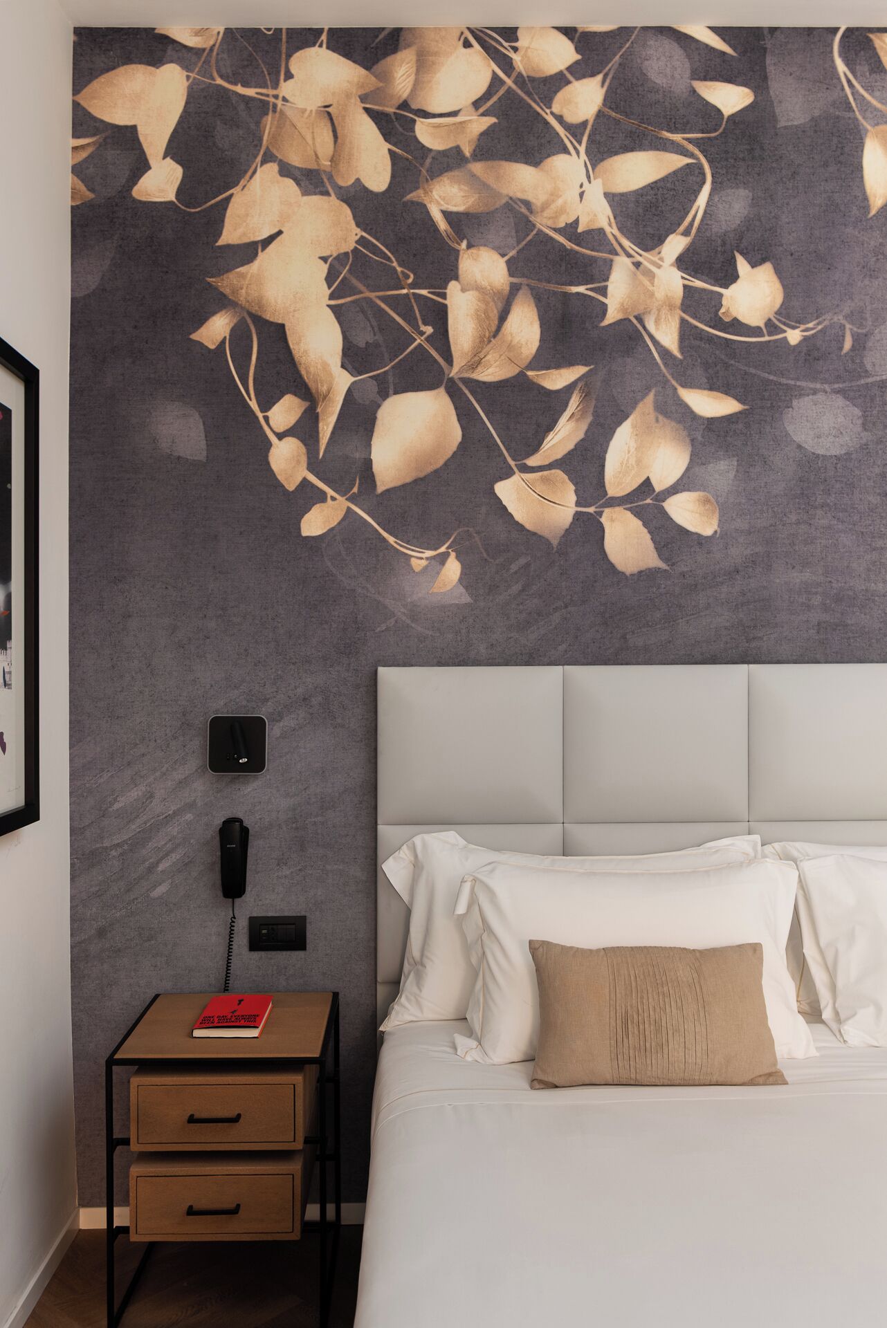 A bedroom with a bed, a nightstand, a wall-mounted phone, and a wall art of leaves.