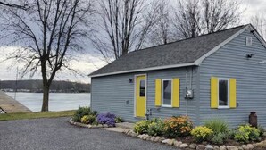 Un piccolo edificio a un piano, con facciata grigia e persiane gialle, circondato da un vialetto sterrato e un giardino curato con arbusti e fiori vari.