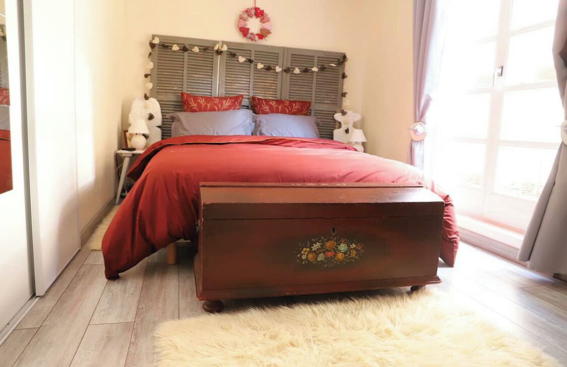 A bedroom with a bed, a dresser, a nightstand, a wall-mounted lamp, and a window with curtains.