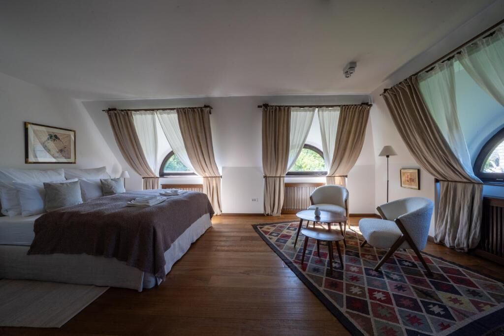 Un dormitorio con cama, dos sillas, una mesa y ventanas amplias con cortinas.