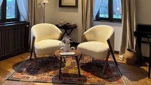 Une pièce avec deux chaises, une petite table, un tableau encadré au mur, un radiateur et une fenêtre avec des rideaux.