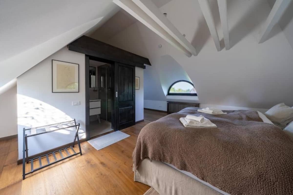 Una camera da letto con soffitto spiovente, un letto con coperta marrone, una finestra e una porta che dà su un'altra stanza.