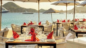Ein Restaurant am Meer mit gedeckten Tischen, großen Sonnenschirmen und Blick auf das Meer und die Schiffe.