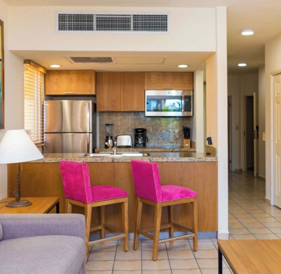 A compact kitchen with wooden cabinets, a stainless steel refrigerator, a microwave, and a coffee maker. There are two pink bar stools and a small table with a lamp.