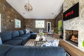 A living room with a stone wall, a fireplace, a sofa set, and a flat-screen TV mounted on the wall. - Historic Brick Retreat w/ Sauna, Pool & Games (Columbus)