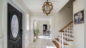 A hallway with a black door, white walls, a chandelier, a staircase, and a dining area in the background. - Historic Brick Retreat w/ Pool & Games (Columbus)