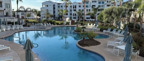 Un área de piscina con sillones de descanso y sombrillas, rodeada de palmeras y edificios.