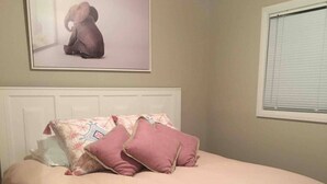 Une chambre à coucher avec un lit, des oreillers, une couverture, une table de chevet, un tiroir, une fenêtre et un tableau au mur.