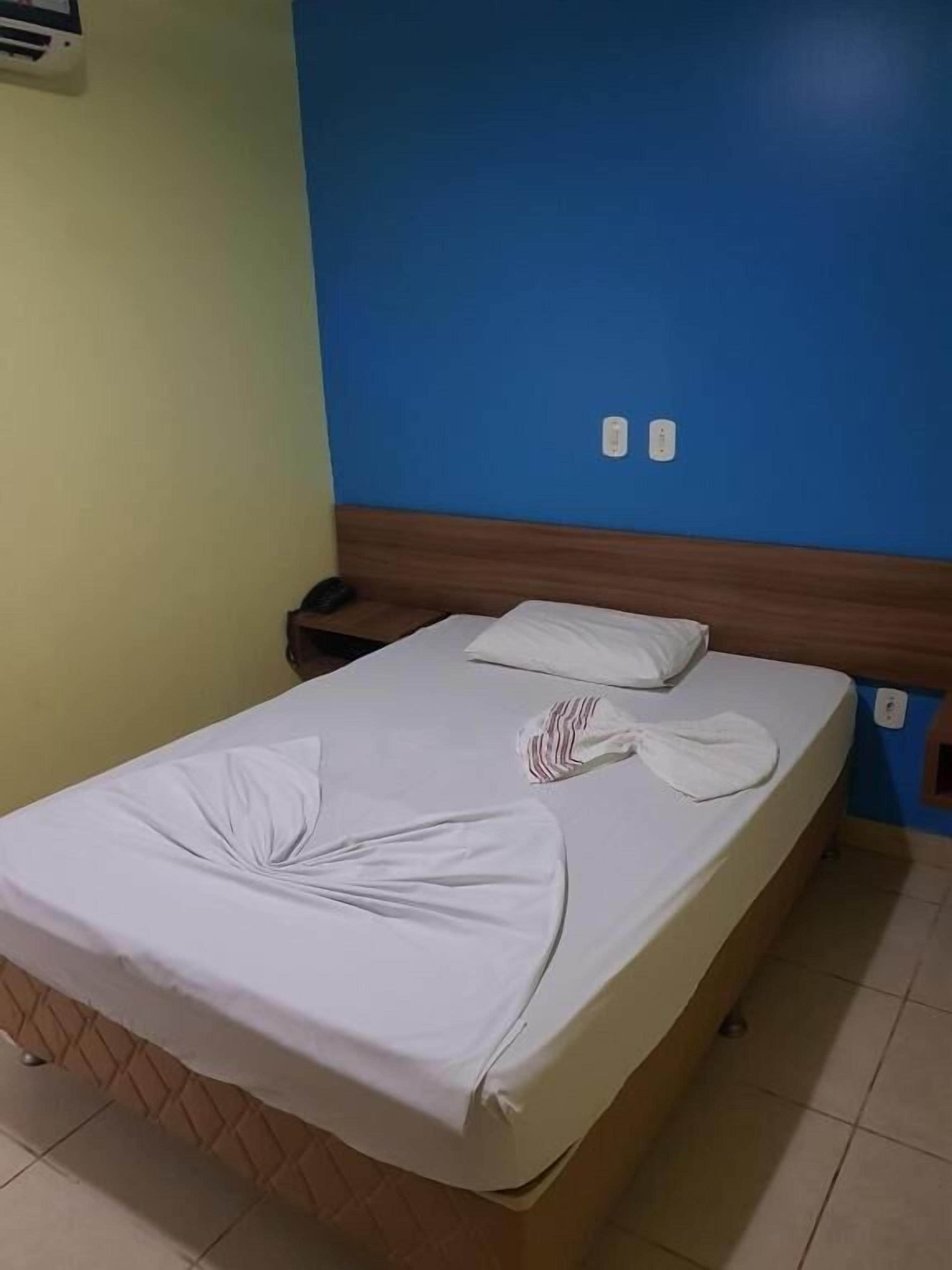Una cama individual con cabecera de madera, una colcha blanca y una toalla blanca doblada con cuidado.
