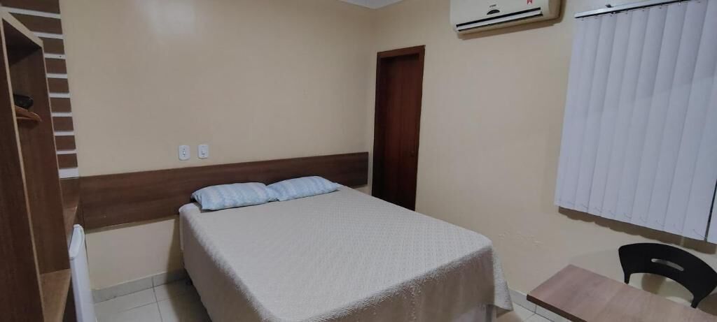 A bedroom with a bed, a wooden headboard, a desk, a chair, and a window with blinds.