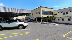 A parking lot with a white SUV, a building with multiple air conditioning units, and a covered area. - Hotel do Porto (Porto Velho)