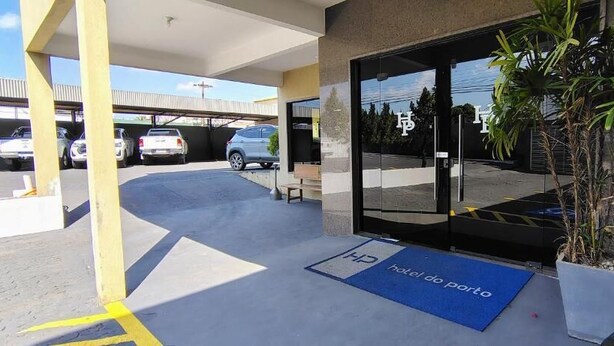 A hotel entrance with a covered parking area, a blue mat with 'Hotel do Porto' text, and a potted plant near the entrance. - Hotel do Porto (Porto Velho)