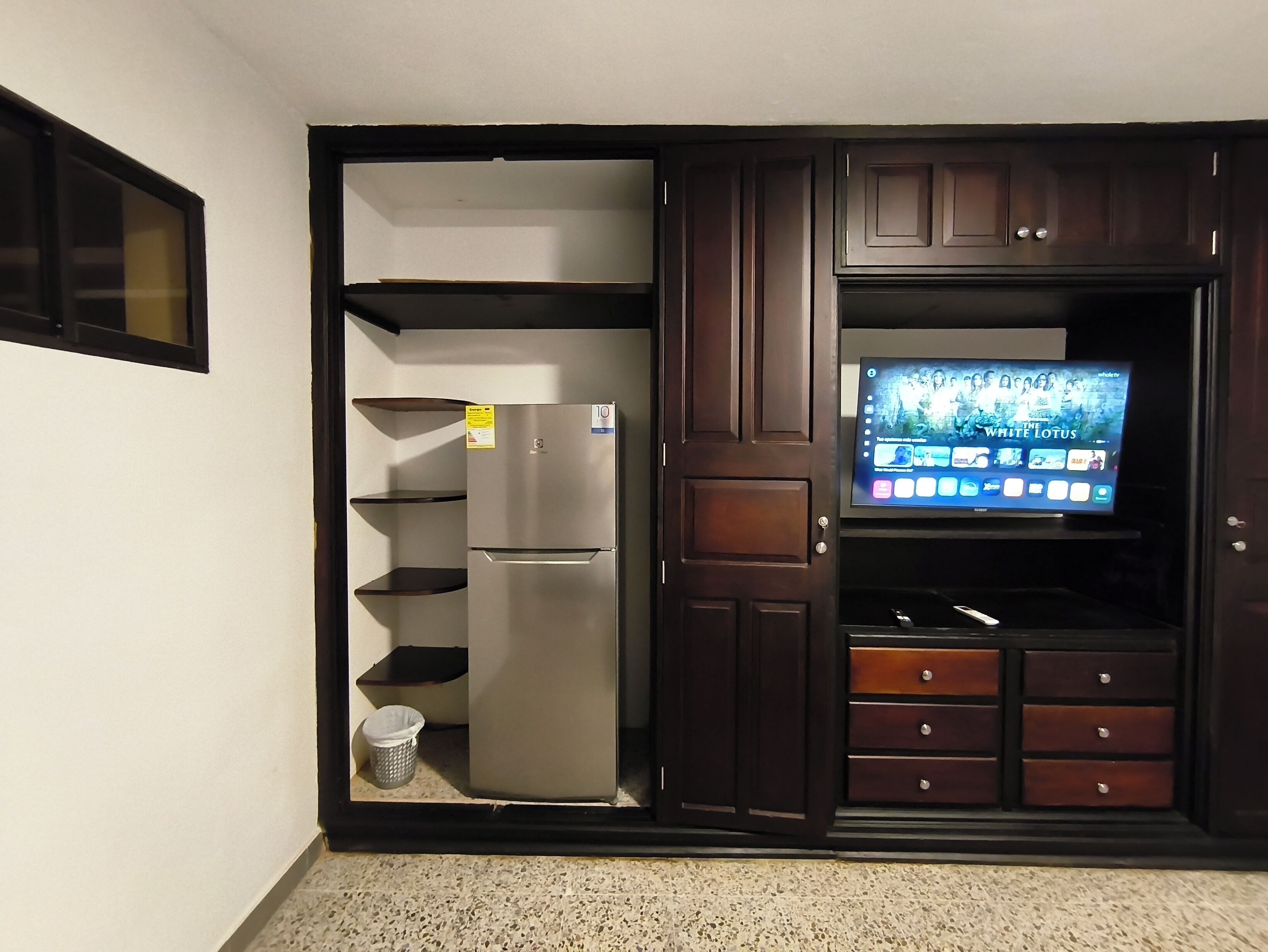 A kitchen with a refrigerator, a TV mounted on a cabinet, and a trash can.