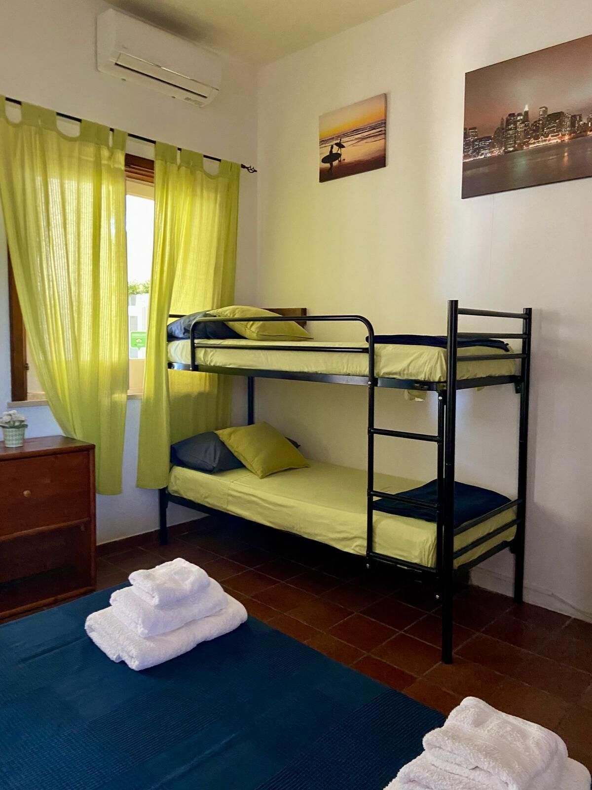 Une chambre avec un lit superposé, une commode en bois, une fenêtre avec des rideaux verts et un tableau représentant un paysage urbain accroché au mur.