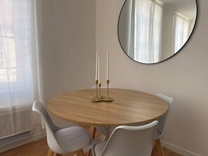 Ein runder Holztisch mit zwei weißen Stühlen, ein Spiegel an der Wand und ein Fenster mit transparenten Vorhängen.