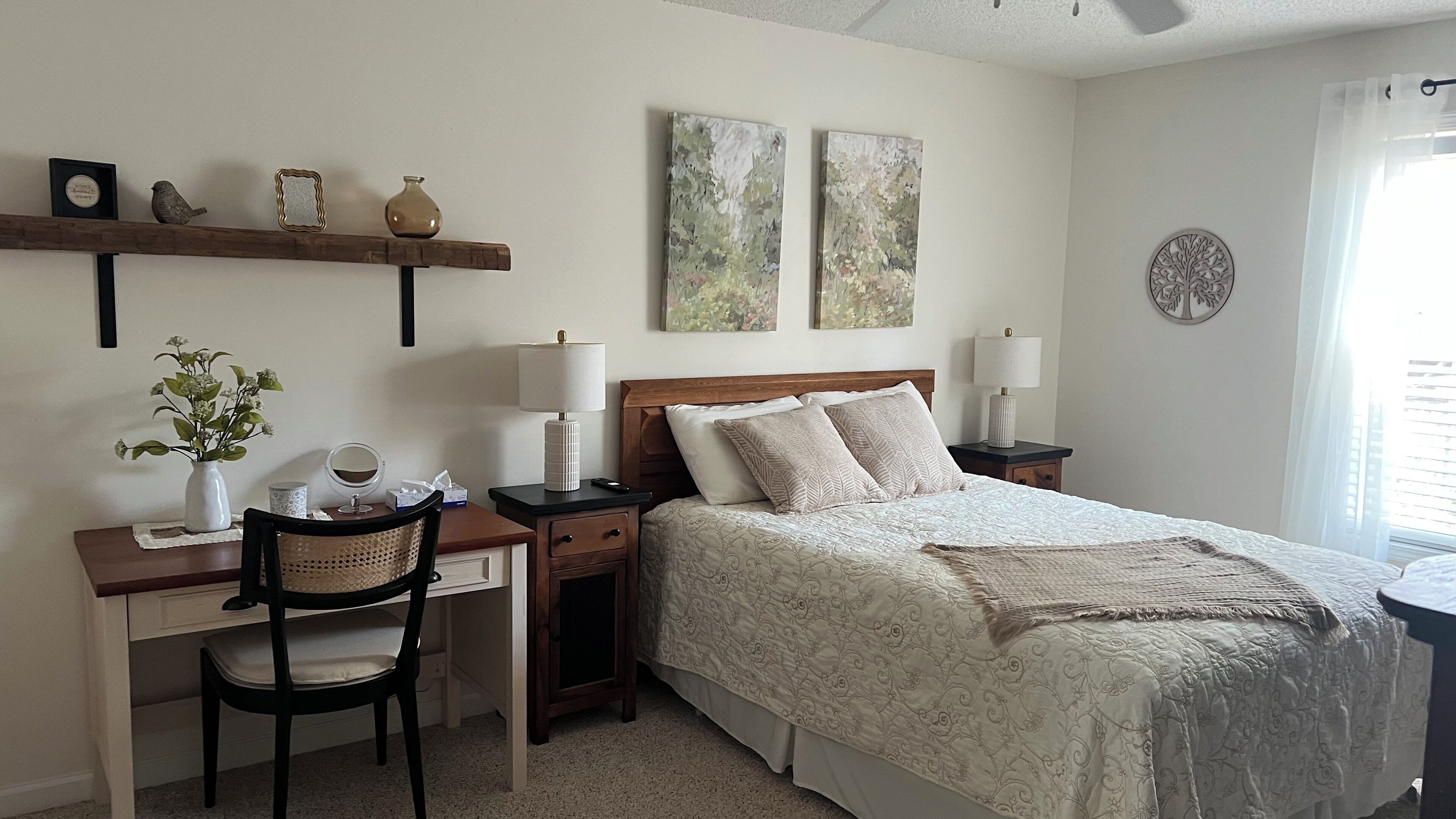 A bedroom with a bed, desk, chair, bedside tables, and wall art.