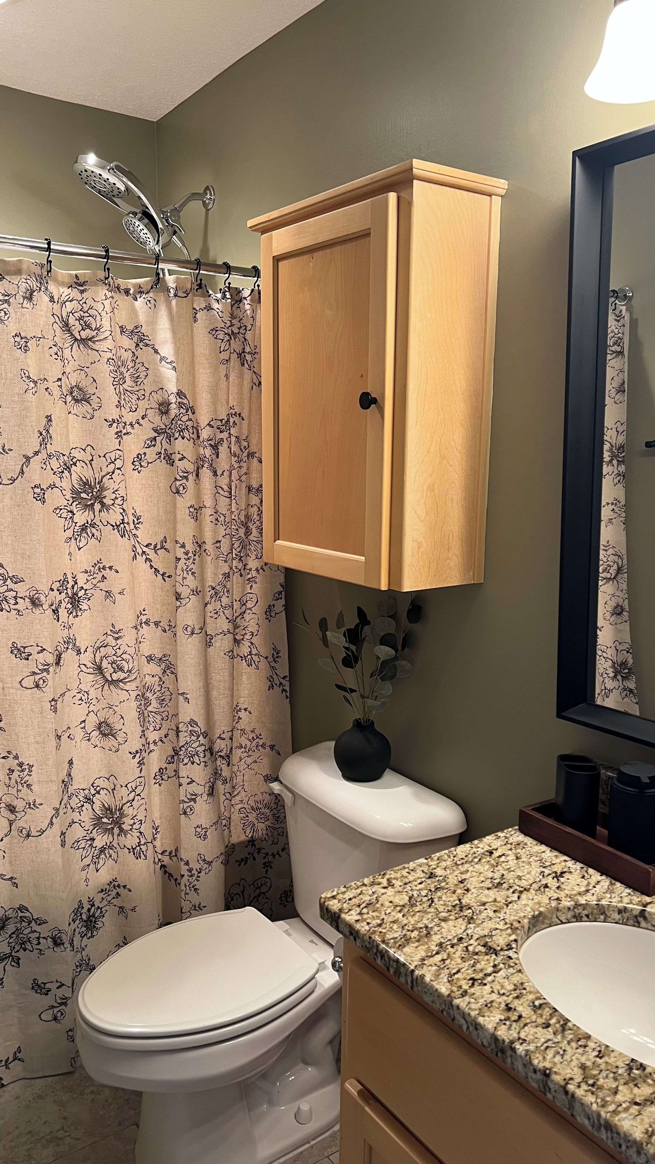 A bathroom with a toilet, a wooden cabinet, a granite countertop, a shower with a floral curtain, and a vase with a plant on the toilet.
