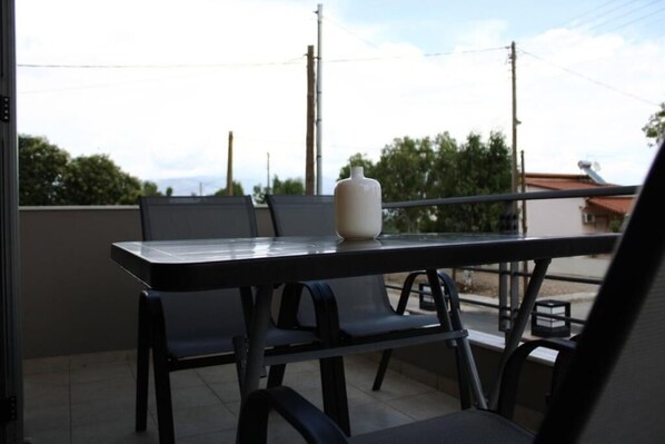 A balcony with a table and chairs, a white vase, and a view of trees and buildings. - Dream (Aigialeia)