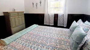 Una camera da letto con un letto, una ventola a soffitto, una finestra con tende leggere, un comò e decorazioni alle pareti.