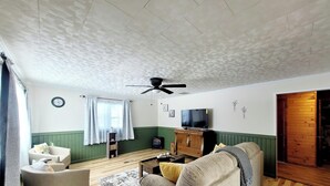 Un soggiorno con ventilatore a soffitto, televisore, divano, tavolino da caffè e finestra con tende.