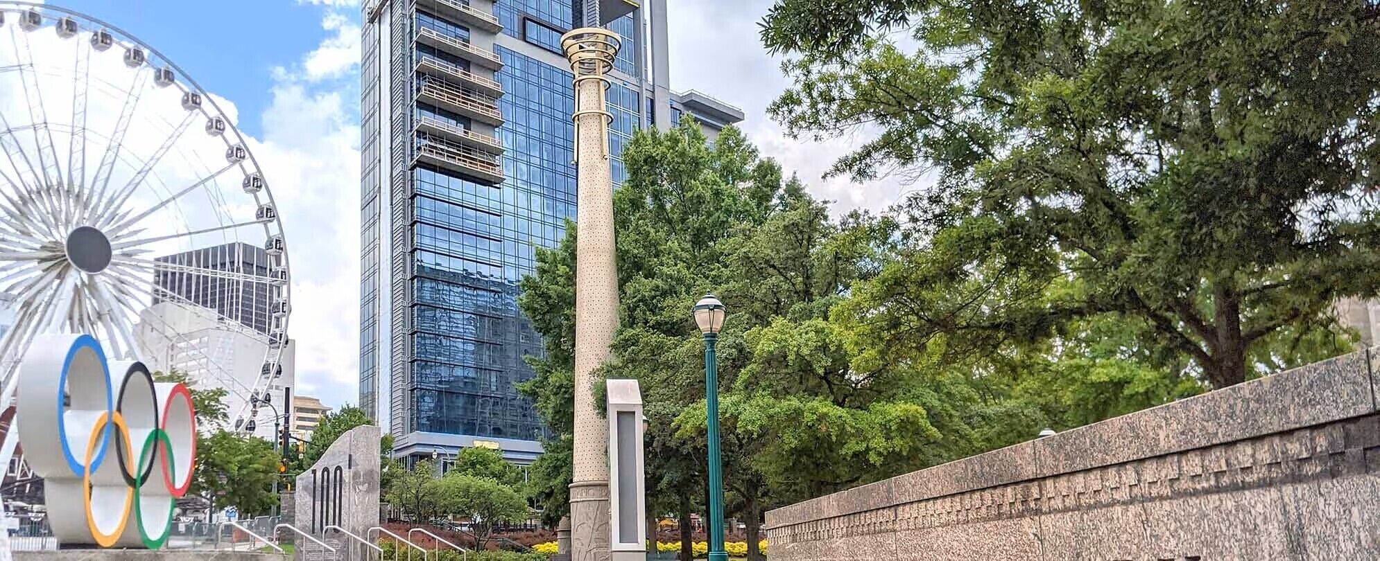 Una ruota panoramica, un edificio moderno in vetro, un'alta colonna e un muro di pietra con alberi sullo sfondo.