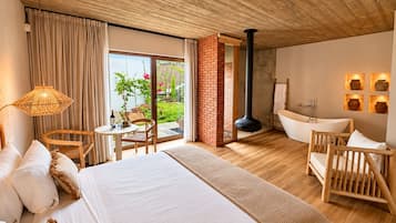 A hotel room with a large bed, a sitting area with a table and chairs, a bathtub, and a view of the garden.