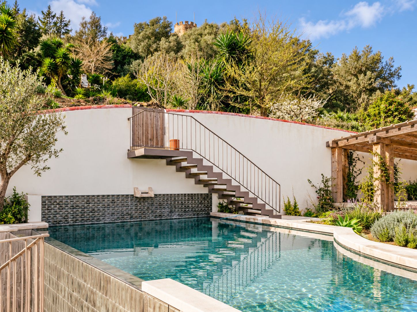 A swimming pool with a wooden deck, a staircase leading to a balcony, and a view of a castle in the background.