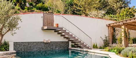 Une piscine avec une terrasse en bois, un escalier menant à un balcon et, au loin, une vue sur un château.