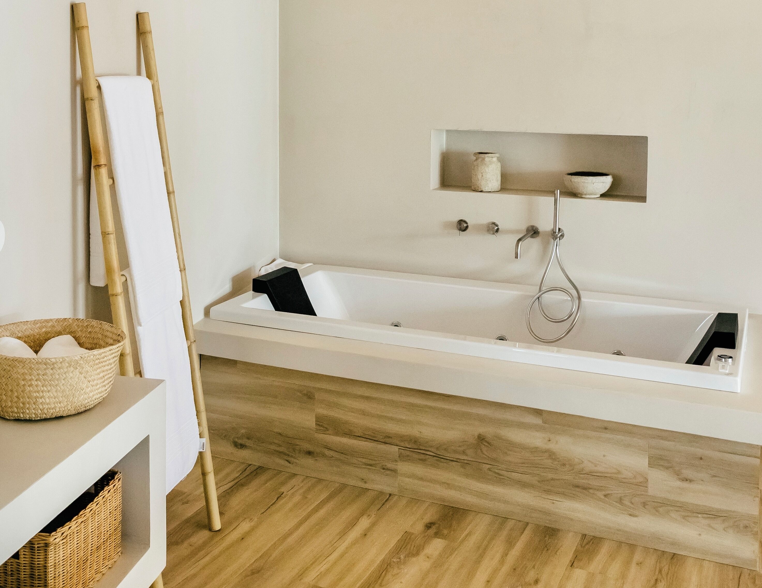 Uma casa de banho moderna com soalho de madeira, uma banheira branca, uma escada de bambu e uma prateleira com frascos e tigelas.