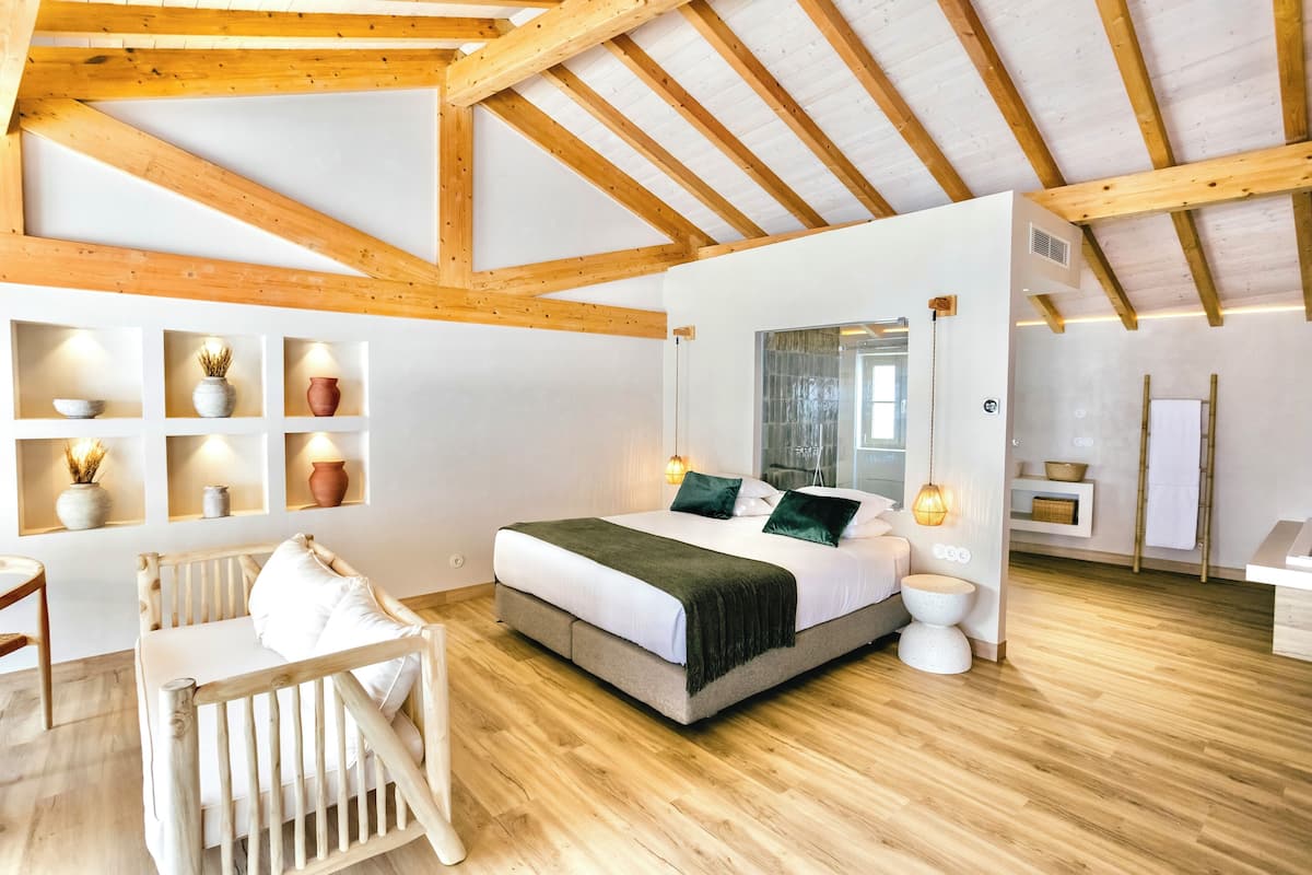 A bedroom with a wooden ceiling, a bed with a white and green bedspread, bedside tables, a white chair, and shelves with decorative items.