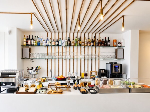 Zona de bar con una variedad de botellas y vasos en estantes, una cafetera y un bufé con alimentos.