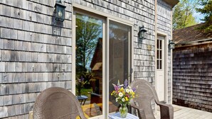 Une façade en bois avec une porte-fenêtre coulissante, des chaises en osier, une petite table ornée d’un vase de fleurs et un numéro de maison bien visible.