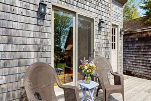 A wooden exterior with a sliding glass door, wicker chairs, a small table with a vase of flowers, and a visible house number. - Mount Desert Island View (Trenton)