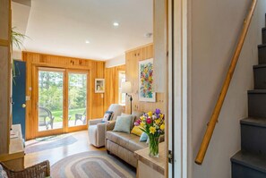 A modern living room with a sofa, a coffee table, and a vase of flowers. There is a staircase to the right and a door leading outside. - Mount Desert Island View (Trenton)