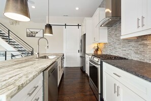 Una cocina moderna con armarios blancos, encimera de mármol, estufa de acero inoxidable y una lámpara colgante.