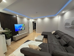 A modern living room with a large flat-screen TV, a dark leather sofa, a white coffee table, and a wooden floor.